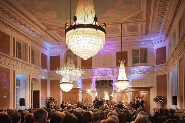 Muzyczny Festiwal w Łańcucie w Sali Balowej, publiczność siedzi, artyści na scenie, świecące żyrandole