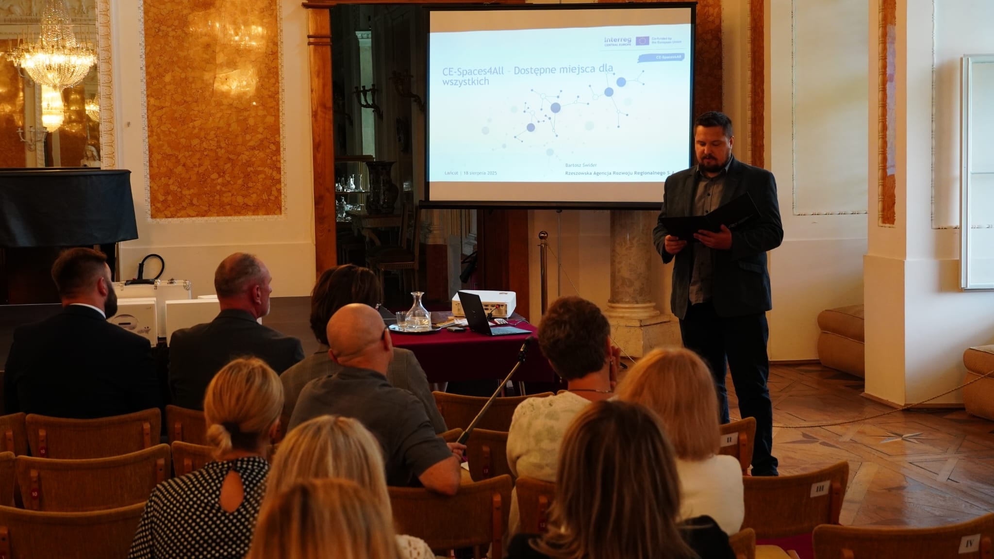 Sala Balowa, Kamil Ulma z MPRŻ mówi do zebranych, stoi. Przed nim siedzący słuchacze. W tle ekran z prezentacją multimedialną.