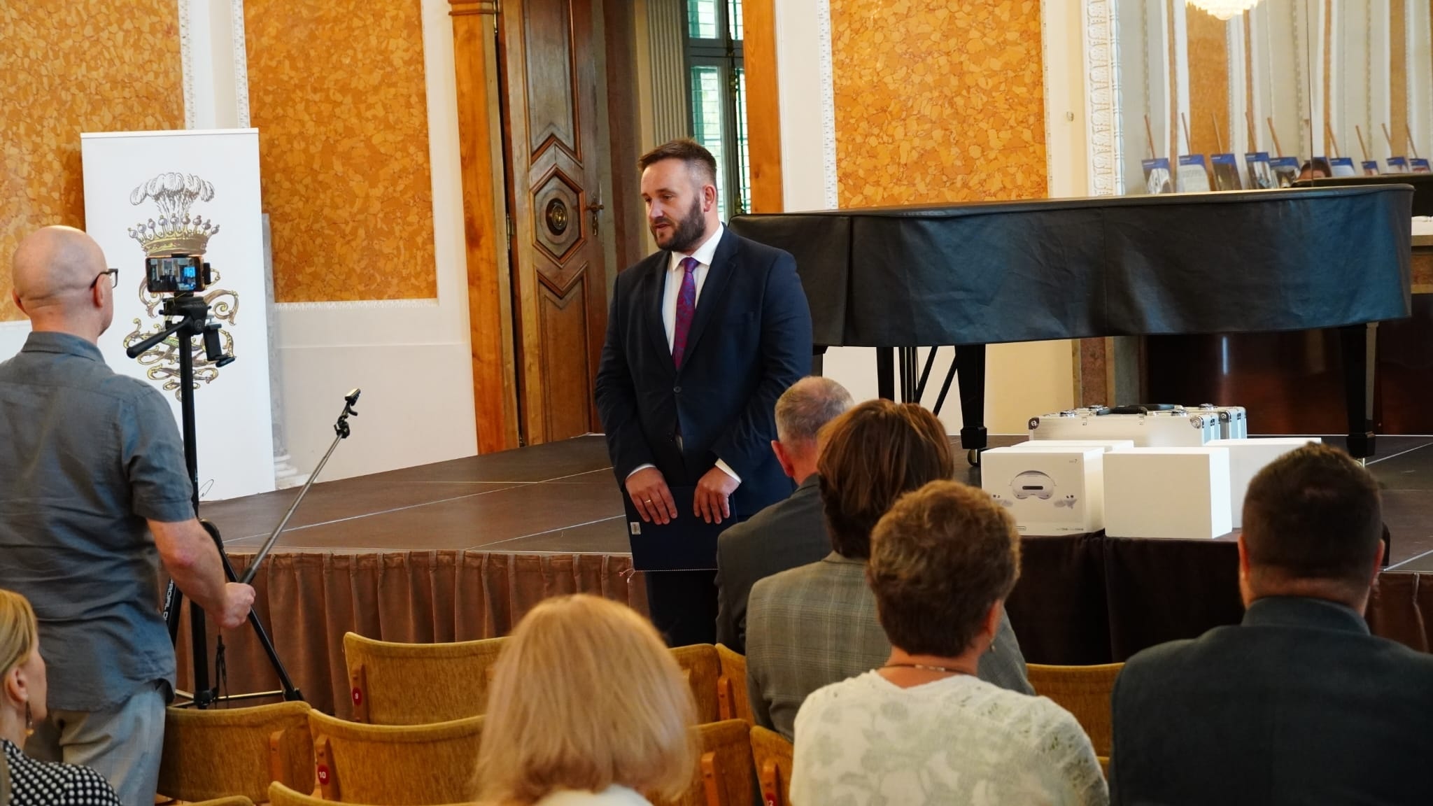 Sala Balowa, dyrektor dr Piotr Szopa mówi do zebranych, stoi. Przed nim siedzący słuchacze. W tle scena z fortepianem.
