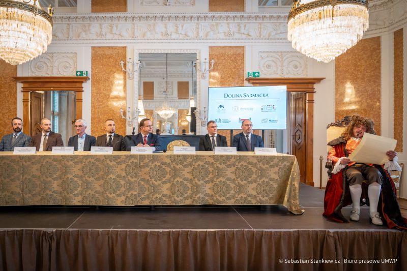 Sala Balowa, aktor w stroju z epoki sarmackiej z odczytem, siedzi, w tle 8 mężczyzn przy stole - uczestnicy podpisujący Dolina Sarmacka