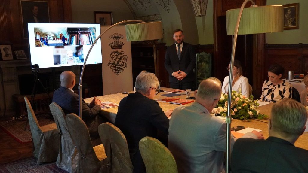Zdjęcie przedstawia posiedzenie Rady Muzeum w zabytkowym wnętrzu Zamku w Łańcucie. Grupa osób siedzi przy długim stole ozdobionym kwiatami. U szczytu stołu stoi przemawiający mężczyzna w garniturze. Po lewej stronie umieszczono duży monitor z widocznym podglądem wideokonferencji oraz baner z herbem zamku. Wnętrze charakteryzuje się historycznym wystrojem, sklepieniami z polichromiami oraz stylowym, ciemnym umeblowaniem. Spotkanie łączy formę stacjonarną z uczestnictwem zdalnym.