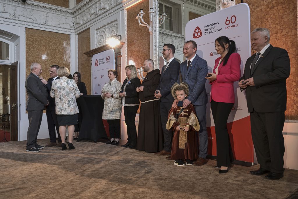 Nadawanie odznaczeń na scenie w Sali Balowej. Odznaczeni stoją w rzędzie obok siebie. Minister wręcza kolejny medal. 