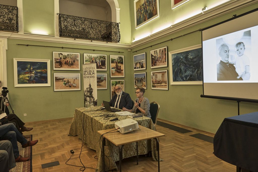 Autorka i prowadzący spotkanie przy stole, autorka mówi do mikrofonu, w tle prezentacja z fotografią A. Dudy-Dziewierza trzymającego na rękach małe dziecko.