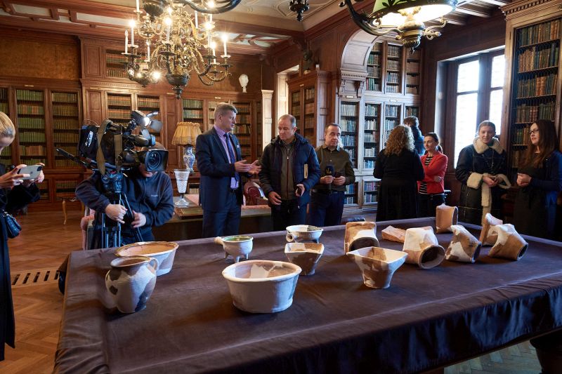 Na pierwszym planie obiekty archeologiczne znalezione w trakcie prac, w tle dziennikarze i uczestnicy rozmawiający po zakończeniu pierwszej części konferencji