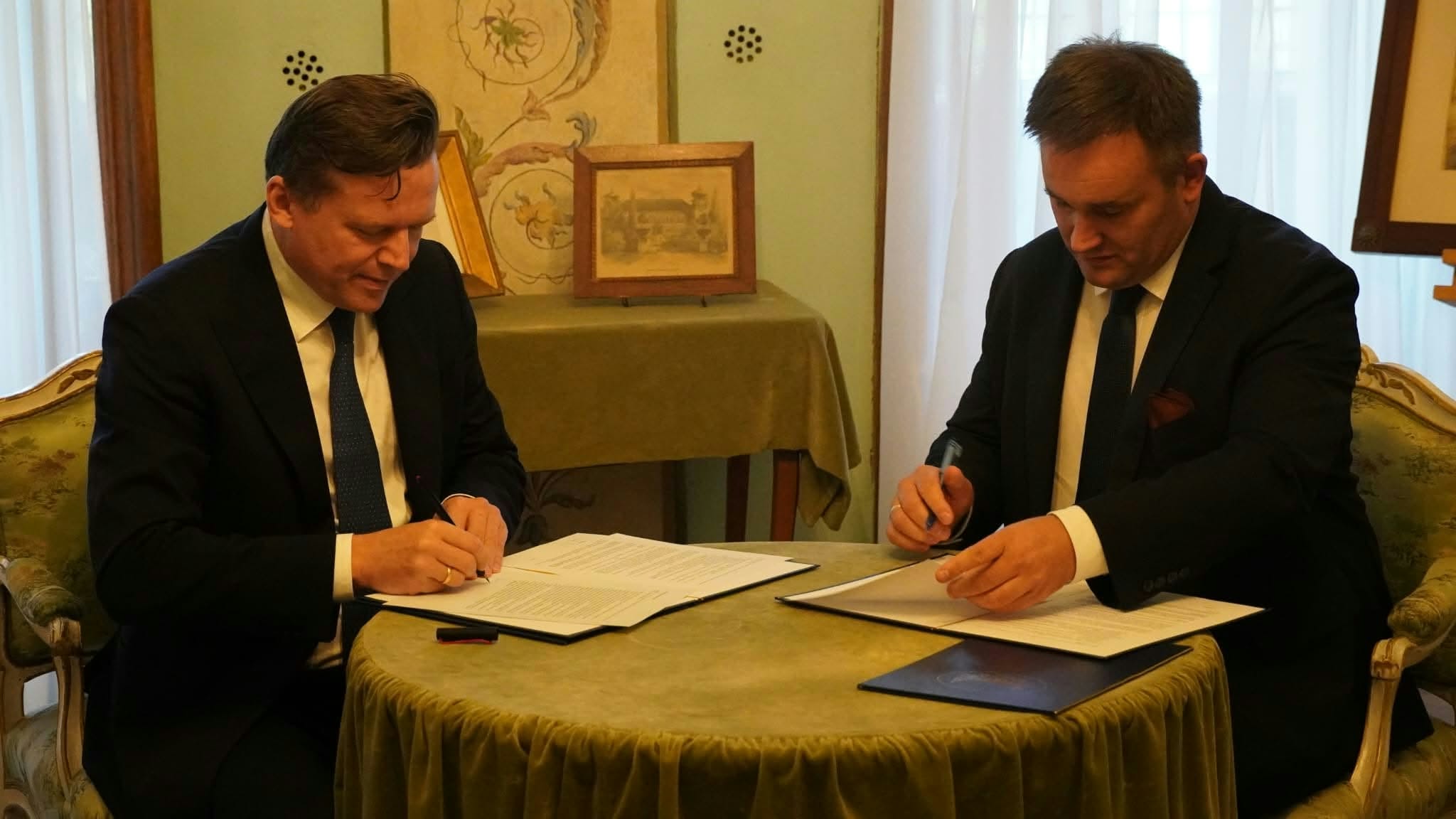 Zdjęcie przedstawia dwóch elegancko ubranych mężczyzn w garniturach, którzy siedzą przy okrągłym, nakrytym zielonym materiałem stoliku i podpisują dokumenty. Mężczyzna po lewej stronie: Siedzi profilem lekko zwrócony w prawą stronę. Ma krótkie, zaczesane do tyłu ciemne włosy, jasną koszulę oraz ciemny krawat w drobne wzory. Pochyla się nad kartkami papieru rozłożonymi na stole i skupiony pisze piórem. Mężczyzna po prawej stronie: Siedzi naprzeciwko, zwrócony lekko w lewą stronę. Ma krótkie, ciemne włosy, jasną koszulę, ciemny krawat oraz garnitur, w którego klapę wpięto małą poszetkę. Prawą ręką trzyma długopis i podpisuje dokument, a lewą przytrzymuje kartkę. Otoczenie: Spotkanie odbywa się w klasycznym wnętrzu. Obaj mężczyźni siedzą na stylizowanych krzesłach z tapicerowanymi oparciami w jasnych, oliwkowo-beżowych odcieniach, ozdobionych rzeźbionym drewnem. W tle, pod jasną ścianą z ozdobnymi motywami, stoi niewielki stolik nakryty obrusem, na którym ustawiono oprawione w ramki obrazy lub grafiki. Po bokach widać okna przesłonięte jasnymi, przezroczystymi zasłonami, przez które sączy się dzienne światło. Całość kompozycji ma formalny i uroczysty charakter.