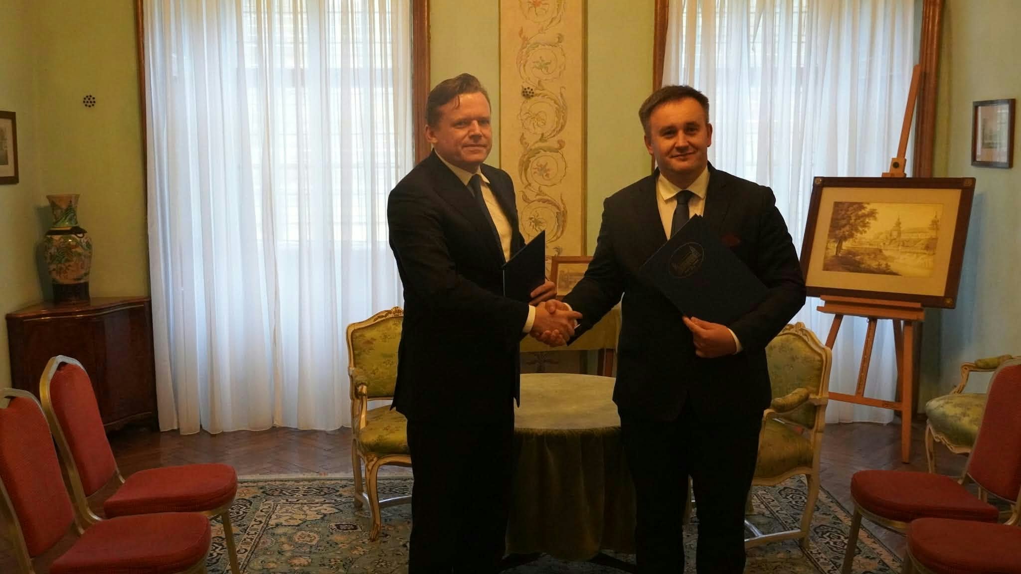 Na zdjęciu widocznych jest dwóch elegancko ubranych mężczyzn w garniturach, stojących w klasycznie urządzonej sali i ściskających sobie dłonie. Mężczyzna po lewej stronie trzyma w drugiej ręce ciemną teczkę, a mężczyzna po prawej przytrzymuje niebieską teczkę z dokumentami. Obaj patrzą w stronę aparatu z uśmiechem. W tle znajduje się okrągły stolik nakryty zielonym obrusem oraz stylizowane krzesła z jasną tapicerką. W pokoju stoją również inne krzesła z czerwonym obiciem, a pod ścianą na sztalugie wyeksponowany jest oprawiony w ramę obraz. Okna w tle przesłonięte są jasnymi, zwiewnymi zasłonami, przez które wpada naturalne światło. Całość ma oficjalny, uroczysty charakter.