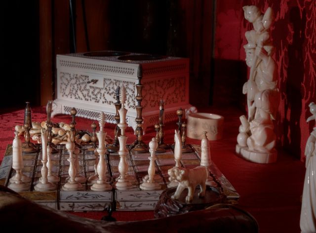 plansza szachowa z pionkami z kości słoniowej, w tle figurki i skrzyneczka. Tło czerwone, z sukna