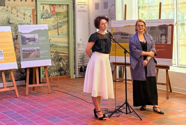 Scena ogólna: Zdjęcie przedstawia wernisaż wystawy w Muzeum – Zamek w Łańcucie. W jasnym pomieszczeniu, na tle fototapety z motywem architektonicznym, stoją dwie kobiety.  Postacie:  Po lewej: Kobieta w okularach, o ciemnych, kręconych włosach, ubrana w czarną bluzkę i białą, plisowaną spódnicę, przemawia do mikrofonu na statywie.  Po prawej: Blondynka w okularach, ubrana w czarne spodnie i wzorzystą, fioletowo-niebieską narzutkę, słucha wypowiedzi z rękami splecionymi przed sobą.  Otoczenie: Po lewej stronie widoczny jest fragment pasiastego, biało-niebieskiego zadaszenia. Za kobietami, na drewnianych sztalugach, stoją tablice informacyjne z tekstem i grafikami (m.in. z napisami „Morskie Wrota Orientu…” i „Indie – Jaipur…”). Po prawej stronie znajduje się duże, półokrągłe okno, przez które wpada jasne światło, a na podłodze ułożona jest czerwona terakota.