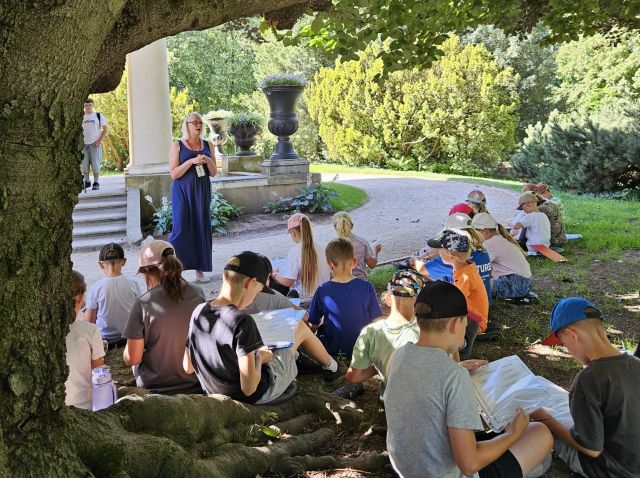 Kadr z zajęć "Lato pod balonem" - kilkanaścioro dzieci siedzi na trawie pod rozłożystym drzewem. Pracownik merytoryczny prowadzi zajęcia. Dzieci mają w dłoniach arkusze papieru i przybory do pisania. Jest ciepły, letni dzień.