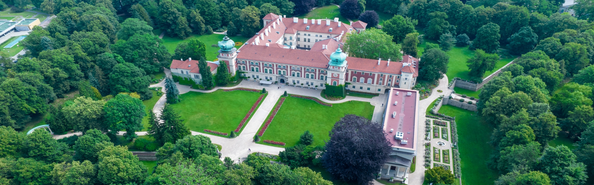 Widok z drona na okazały kompleks zamkowy o jasnych elewacjach z czerwonymi akcentami i mansardowym dachem z czerwonej dachówki. Budowla ma charakterystyczne wieże zwieńczone ozdobnymi, zielonymi hełmami. Przed zamkiem rozciąga się zadbany, duży dziedziniec z równo przystrzyżoną trawą, symetrycznymi alejkami spacerowymi i pasami czerwonych kwiatów. Całość otacza gęsty, zabytkowy park pełen bogatej roślinności i wysokich drzew o różnych odcieniach zieleni, w który wkomponowane są mniejsze obiekty architektoniczne i ogrody kwiatowe.