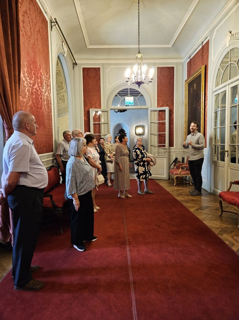 Uczestnicy spotkania - kobiety, mężczyźni zwiedzają ekspozycję I piętra zamku. Po prawej stronie pracownik Muzeum oprowadzający grupę. 
